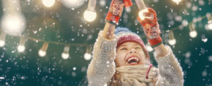 Young girl outside in winter with snow falling. She is laughing as she touches hanging holiday lights.