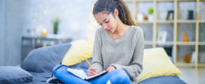 Teen girl sitting on her bed writing in a journal as she reflects on the past year and resents for the new year.