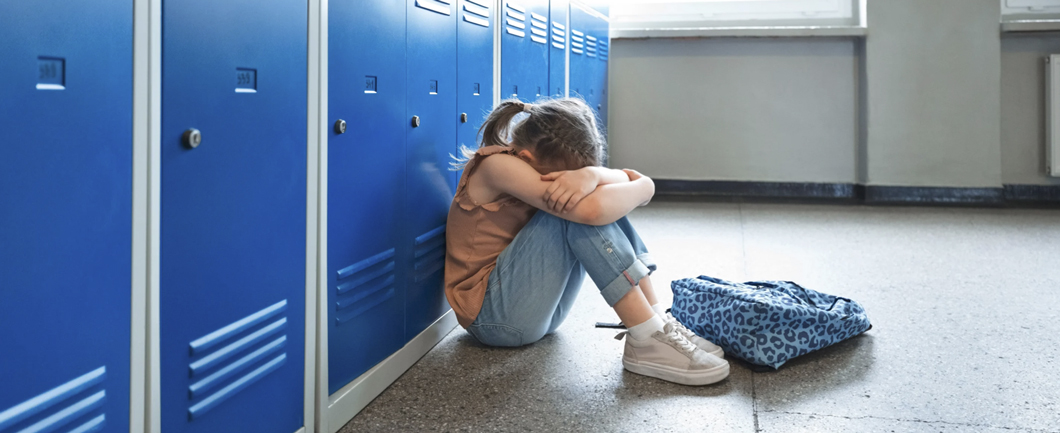Young girl sitting by school lockers with her head down, showing signs of emotional distress.